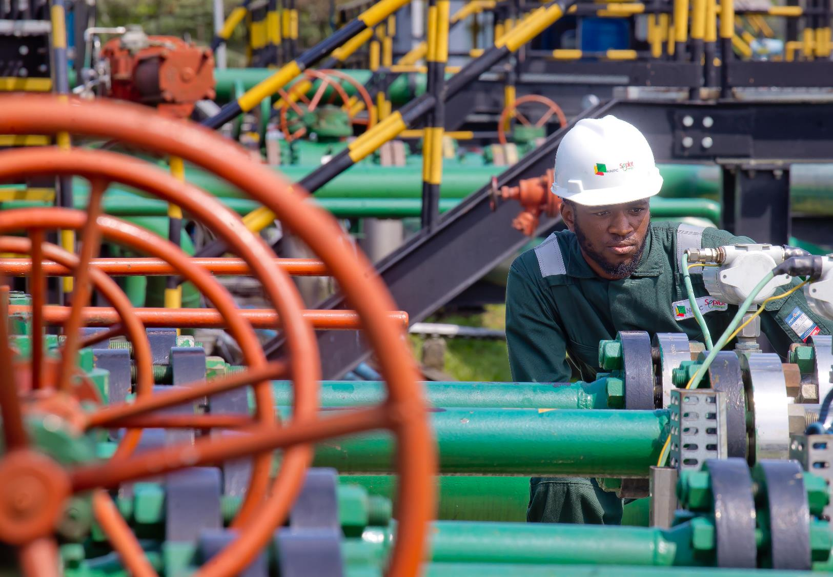 Man in a white hard hat and green coveralls inspecting equipment at an oil field site.