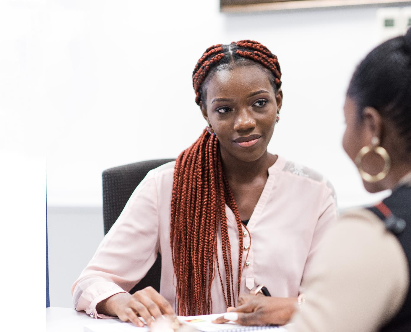 Woman with reddish braided hair in a blush blouse speaking during an office meeting.