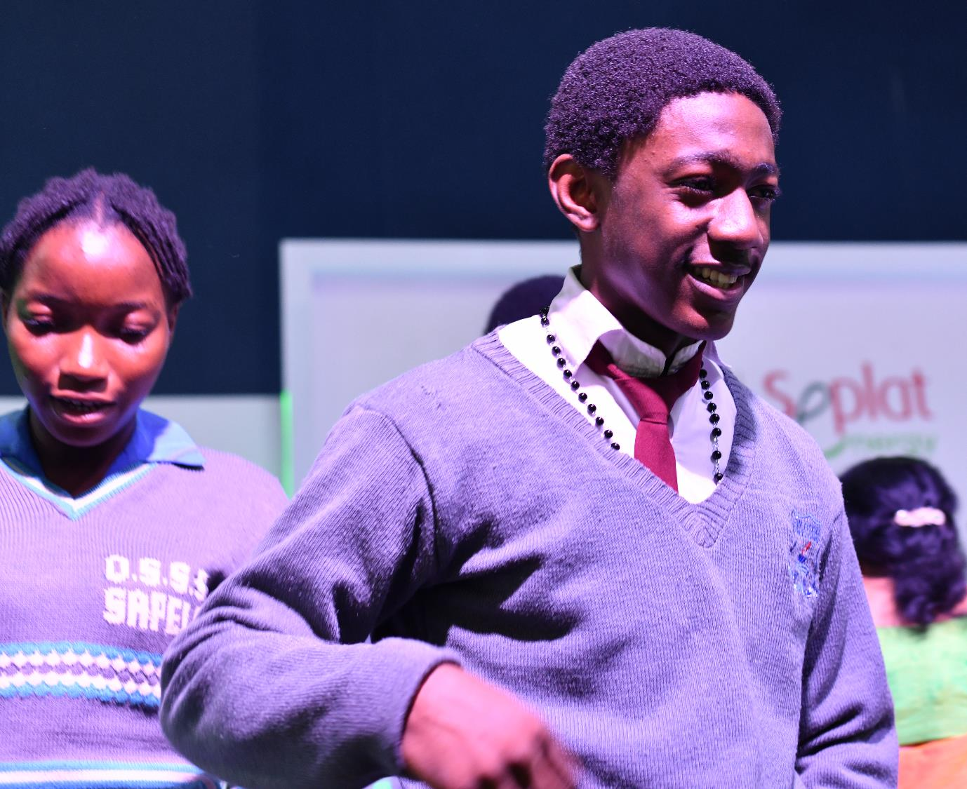 Young man in a grey sweater and red tie smiling in front of a Seplat Energy banner.