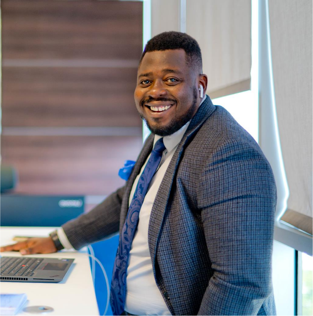 Man in a grey suit and blue tie smiling beside a laptop in an office.